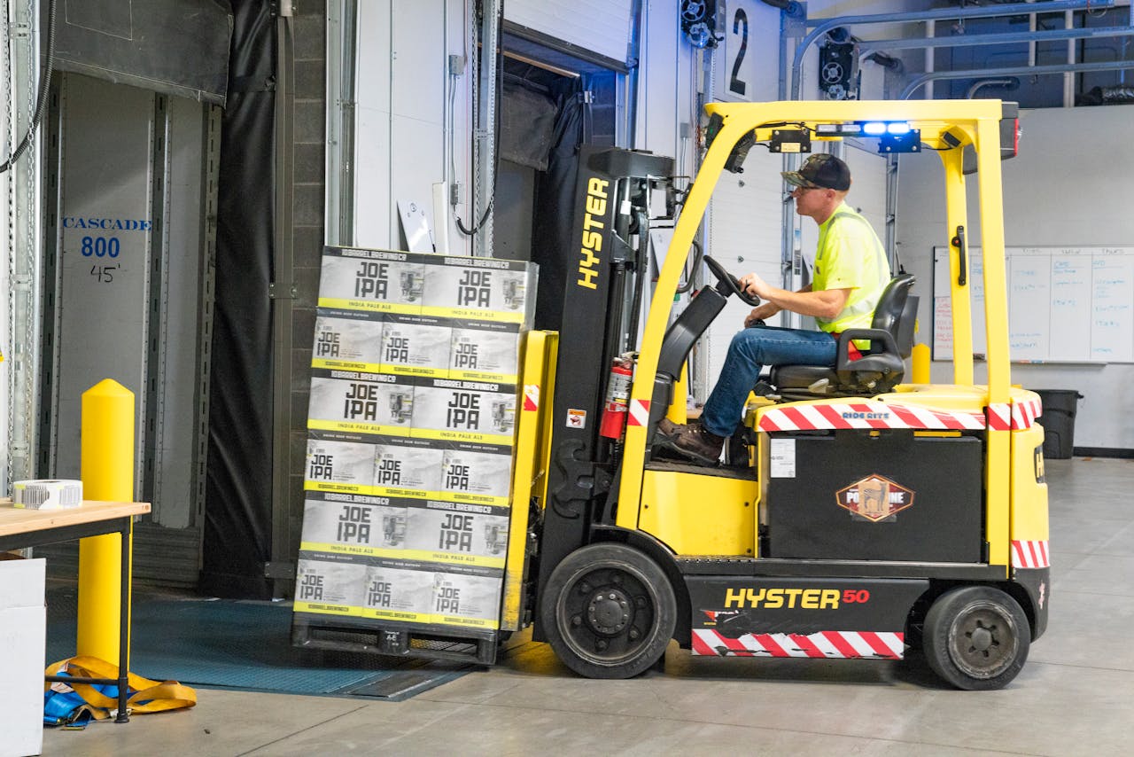 services-02 A worker drives a Hyster forklift moving Joe IPA boxes in a warehouse.